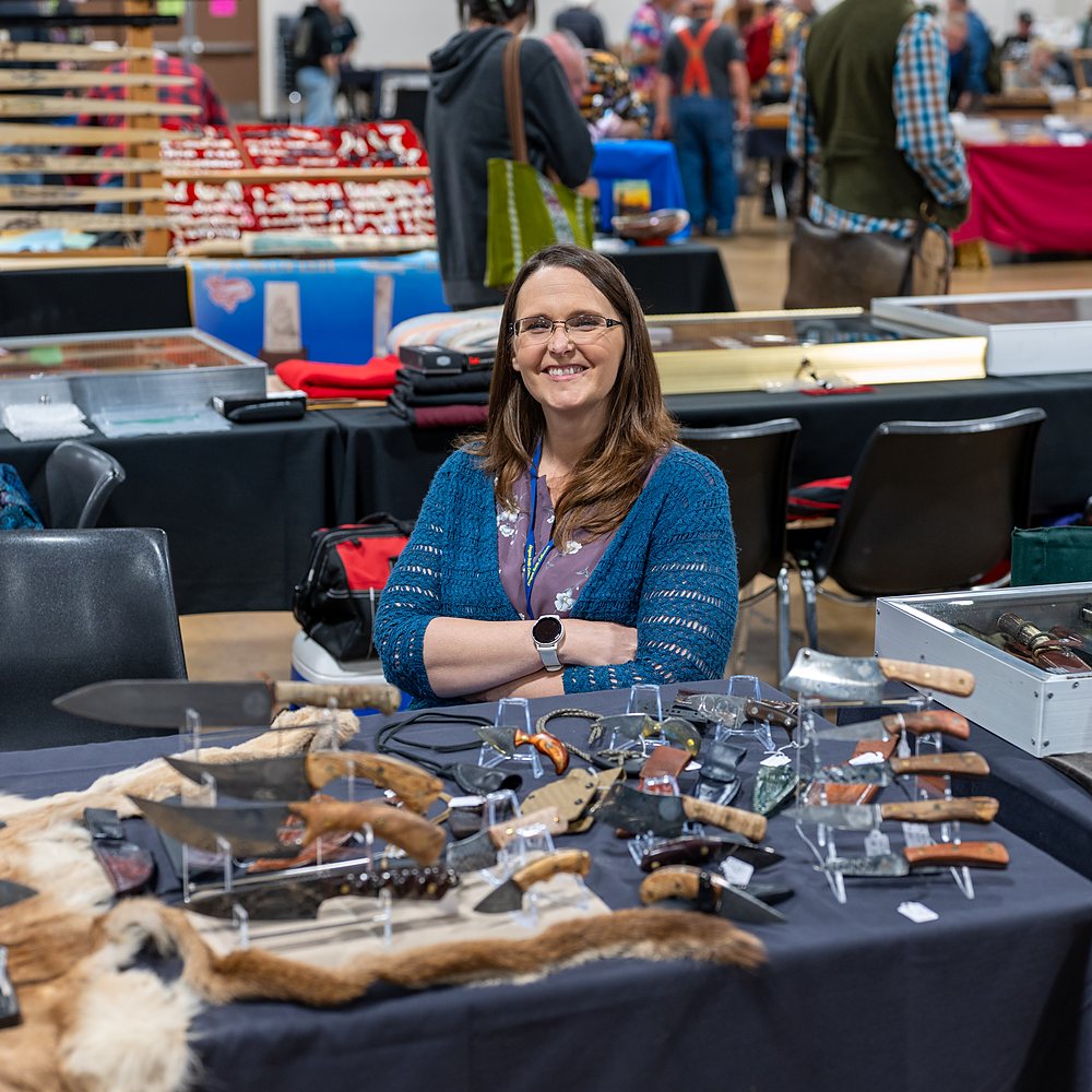 An exhibitor at her table of knives, the hall busy behind her